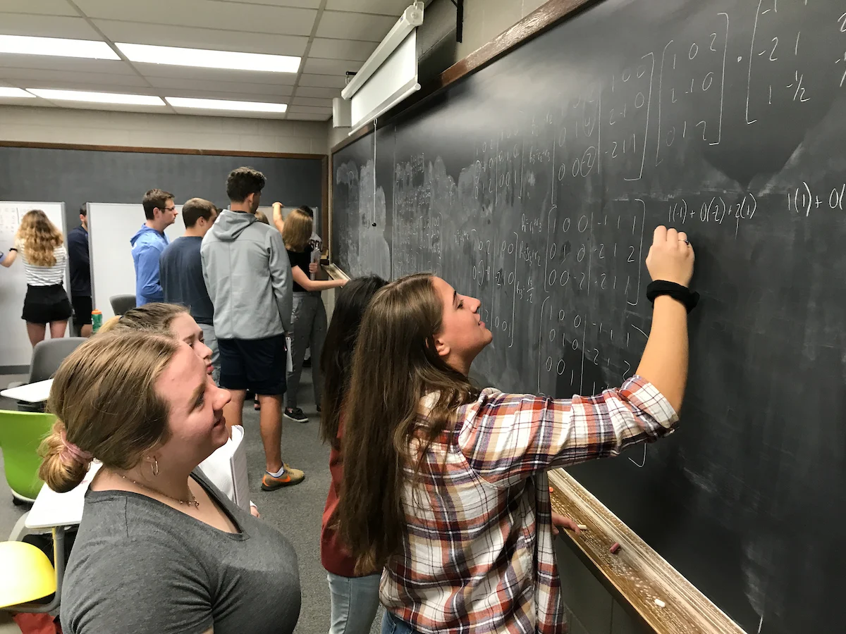 Students in the VERUM Summer Research Program solve math problems together on a blackboard in a classroom, with others watching and participating.