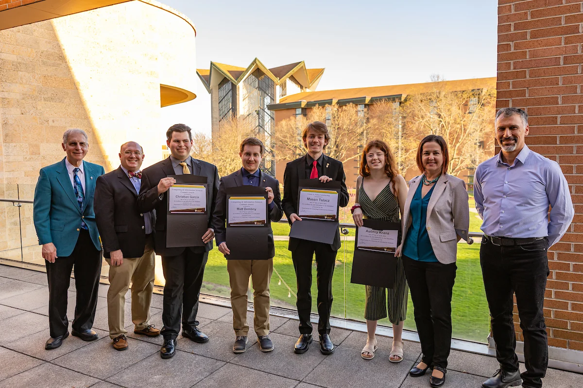 Eight people stand outside; four in the middle hold certificates and folders, smiling for a group photo celebrating their research achievements.