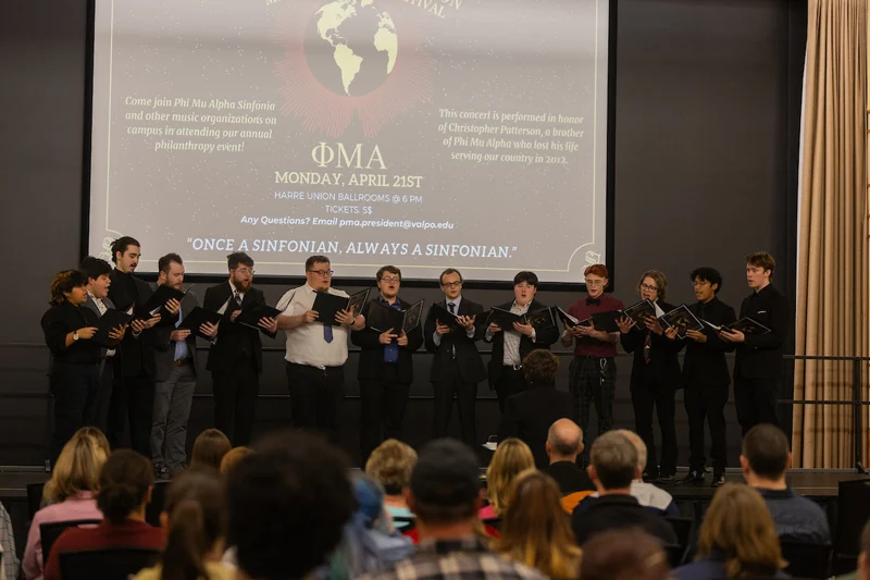 A choir of men in suits sings on stage to an audience, with a large event banner displayed behind them—an inspiring showcase of Music for Non-Majors.