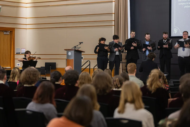 A group of people sing onstage while a violinist plays, captivating the audience seated in front of them. Admission grants you an unforgettable live music experience.