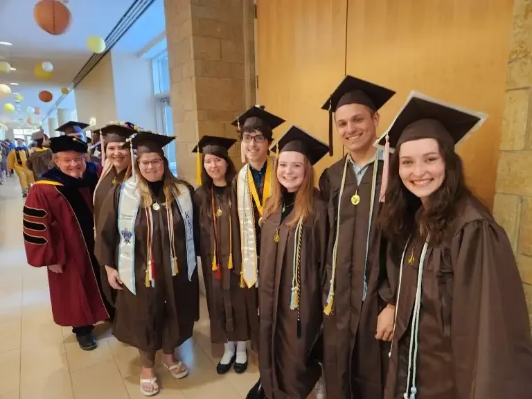 A group of graduates in caps and gowns smiling indoors, with faculty members in academic regalia behind them, celebrating their achievements and successful admission into the next chapter of their lives.