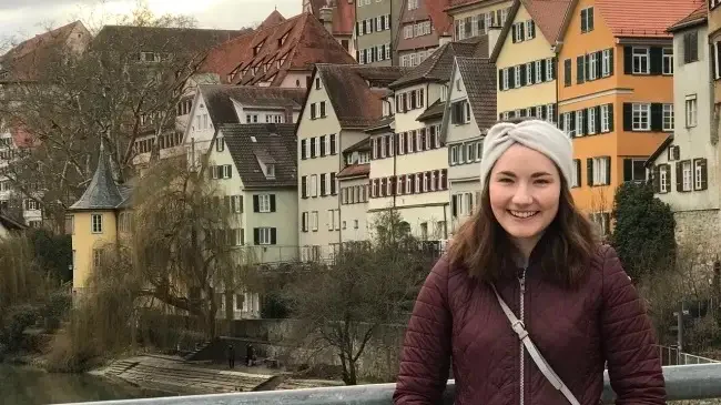 Woman smiling on a bridge with colorful, historic European buildings in the background, capturing the joy of studying abroad.