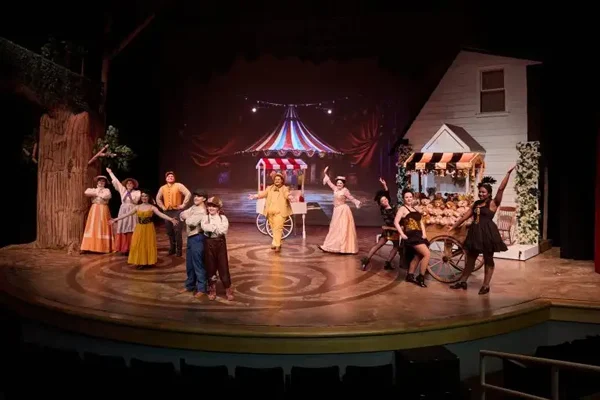 Actors in colorful costumes perform on a stage with a circus tent backdrop and a house set piece, bringing lively energy reminiscent of Music for Non-Majors.