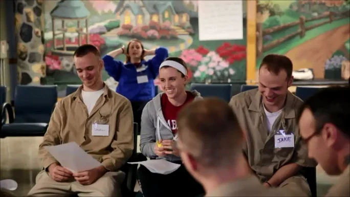 Three people with name tags sit and smile in a circle, engaging in the Inside Out Course, with a mural and a woman posing in the background.