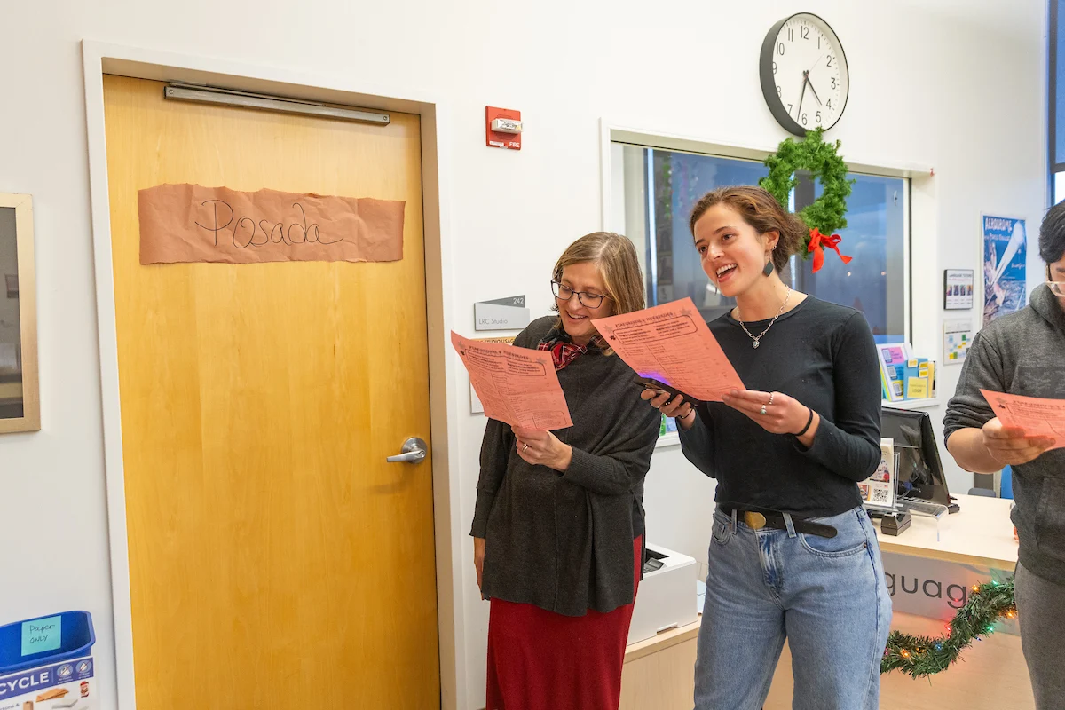 Two women smiling and singing while holding lyric sheets inside an office decorated for the holidays, perfect for student profiles capturing festive campus moments.
