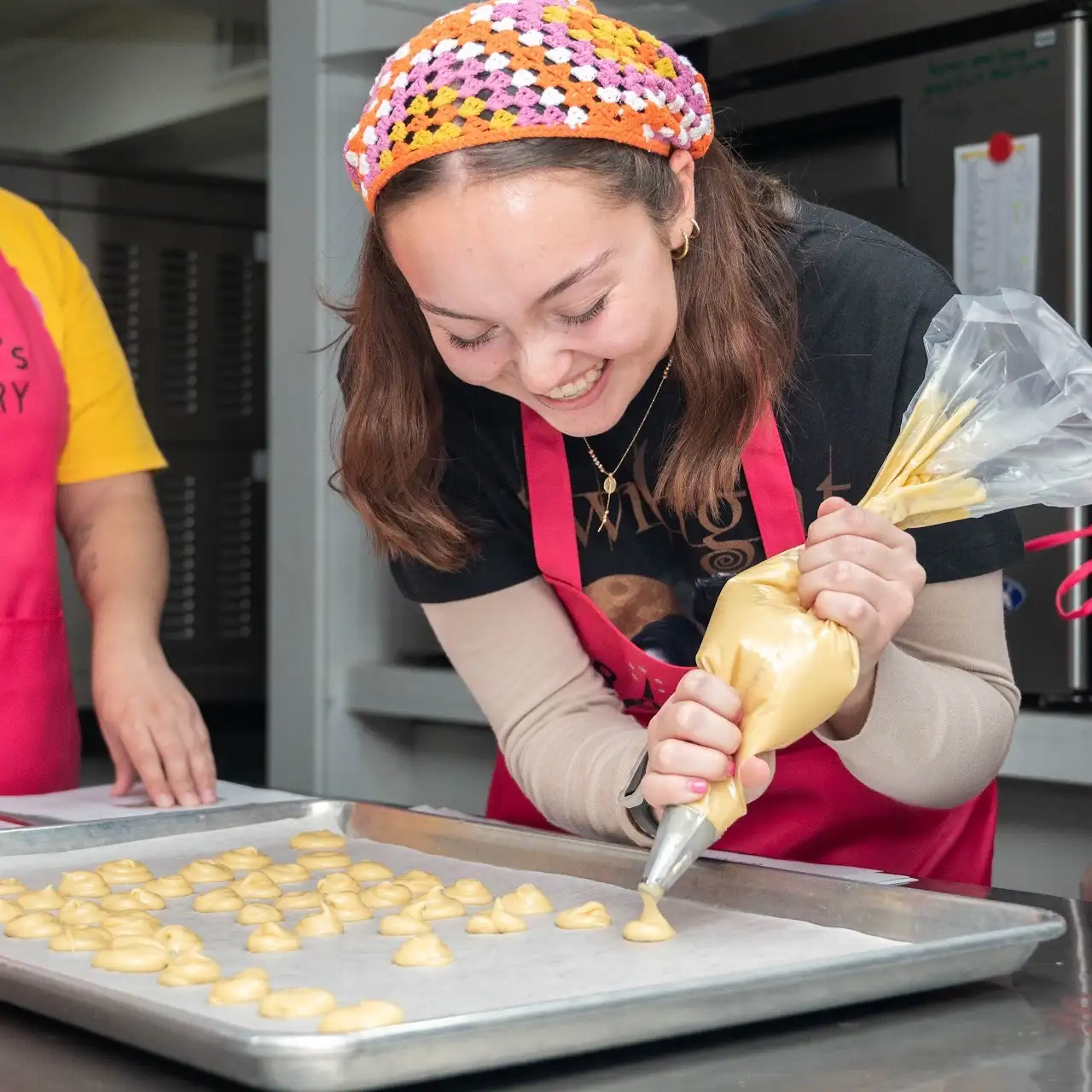 A smiling woman pipes dough onto a baking sheet in a kitchen while wearing a pink apron and colorful headscarf, showcasing student profiles in the world of culinary arts.