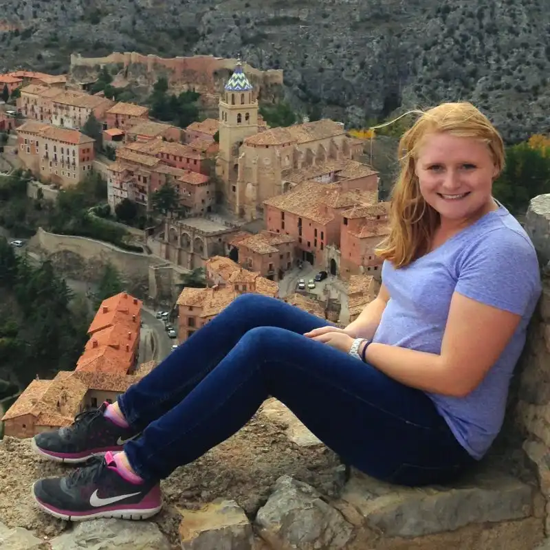 A smiling woman sits on a stone wall overlooking a historic town with terracotta roofs and a church tower, capturing the essence of our Student Profiles.