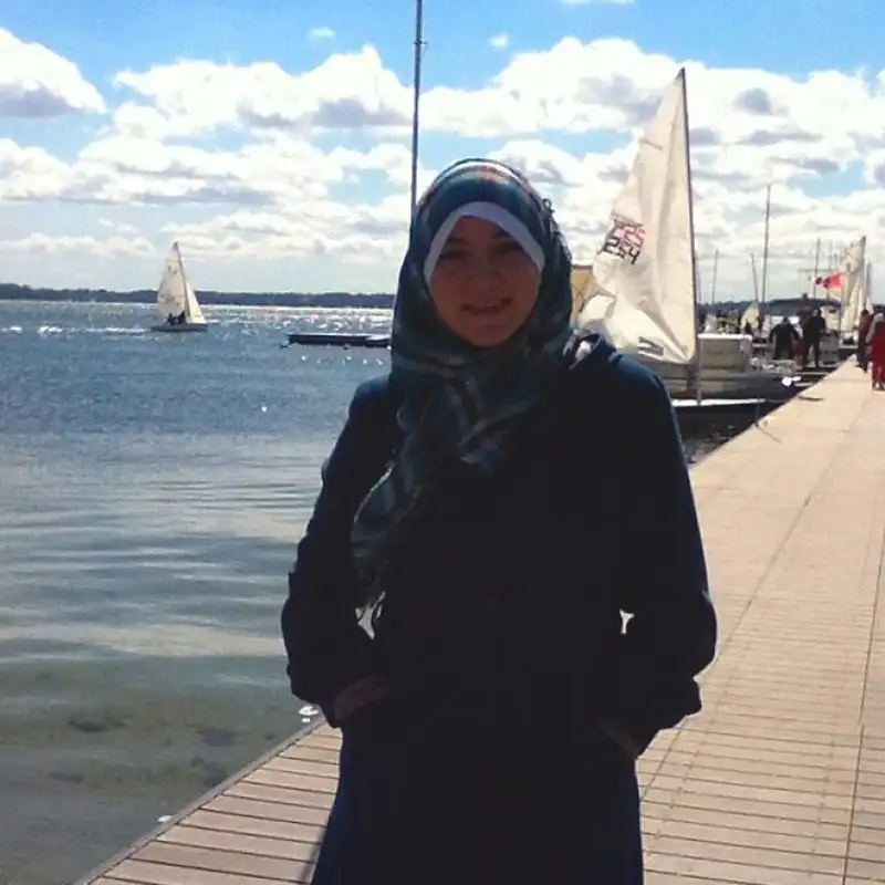 Woman in a hijab stands on a dock by the water, with sailboats and a partly cloudy sky in the background—perfect for Student Profiles capturing campus life.