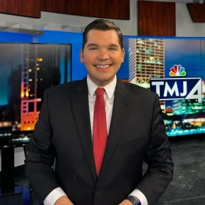 A news anchor, Brendan Johnson, in a black suit and red tie stands smiling in a TV studio with TMJ4 on the screen behind him.