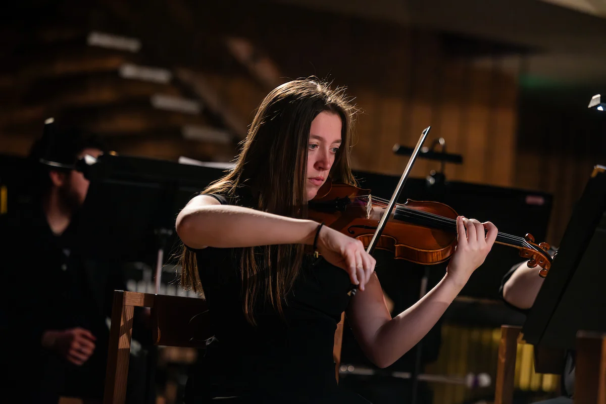 A young woman plays the violin on stage, focused under warm lighting in the background, captivating the audience with her performance as part of an exclusive admission event.