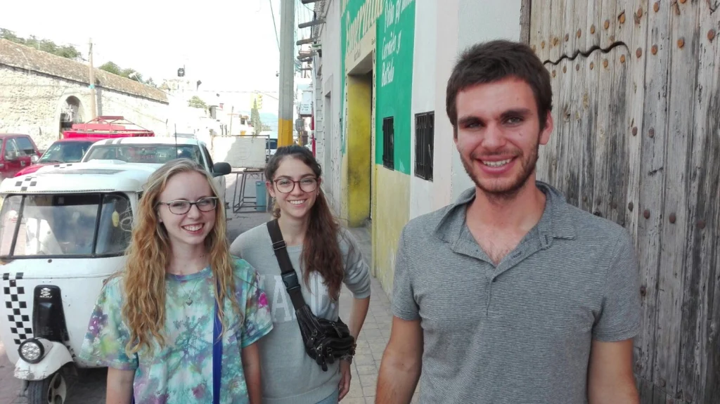 Three smiling young adults stand on a sunny street near a wooden door and a white tuk-tuk.