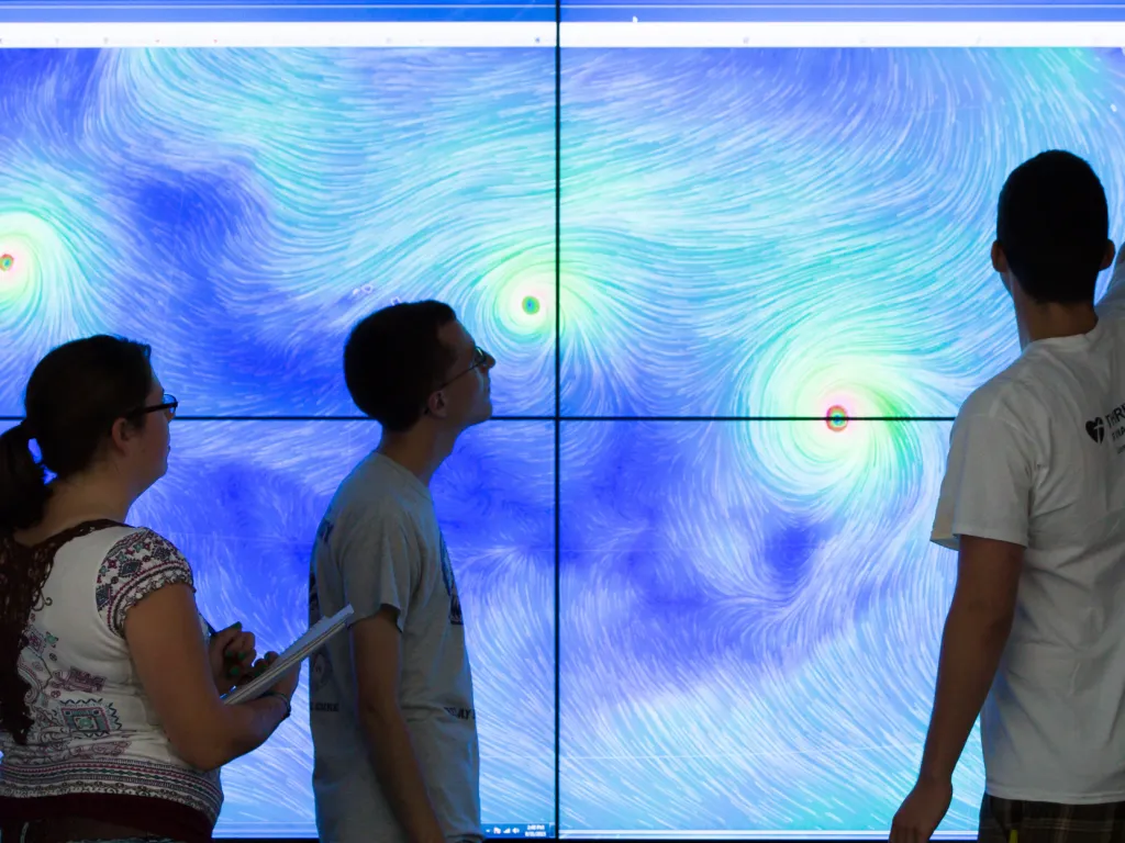 Three people analyze swirling weather patterns displayed on a large screen with blue and green colors.