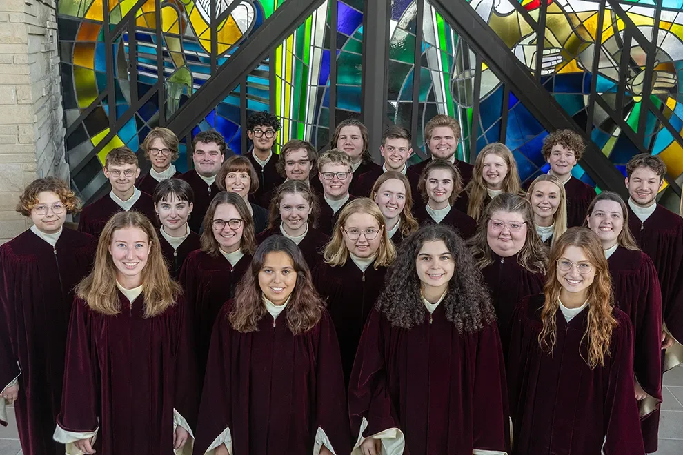 A choir of young adults from the Music Department in maroon robes poses in front of colorful stained glass windows.
