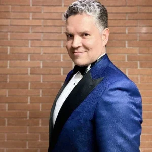 Man from the Music Department in a blue tuxedo smiling, posing in front of a brick wall.