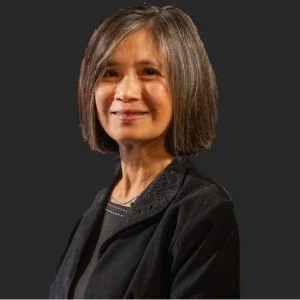 Woman with shoulder-length gray hair wearing a black jacket, smiling against a dark background, representing the Music Department.
