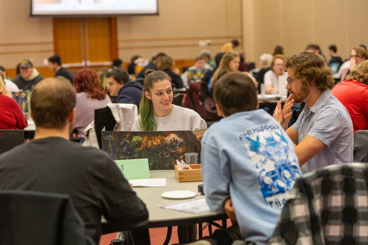 Four people sit around a table playing a tabletop game in a large room filled with Student Organizations and other groups.