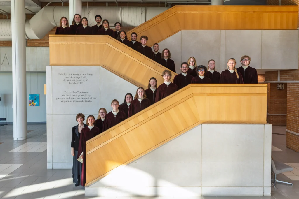 A choir group from Ensemble Tours in matching robes stands on a modern indoor staircase, smiling for a group photo.