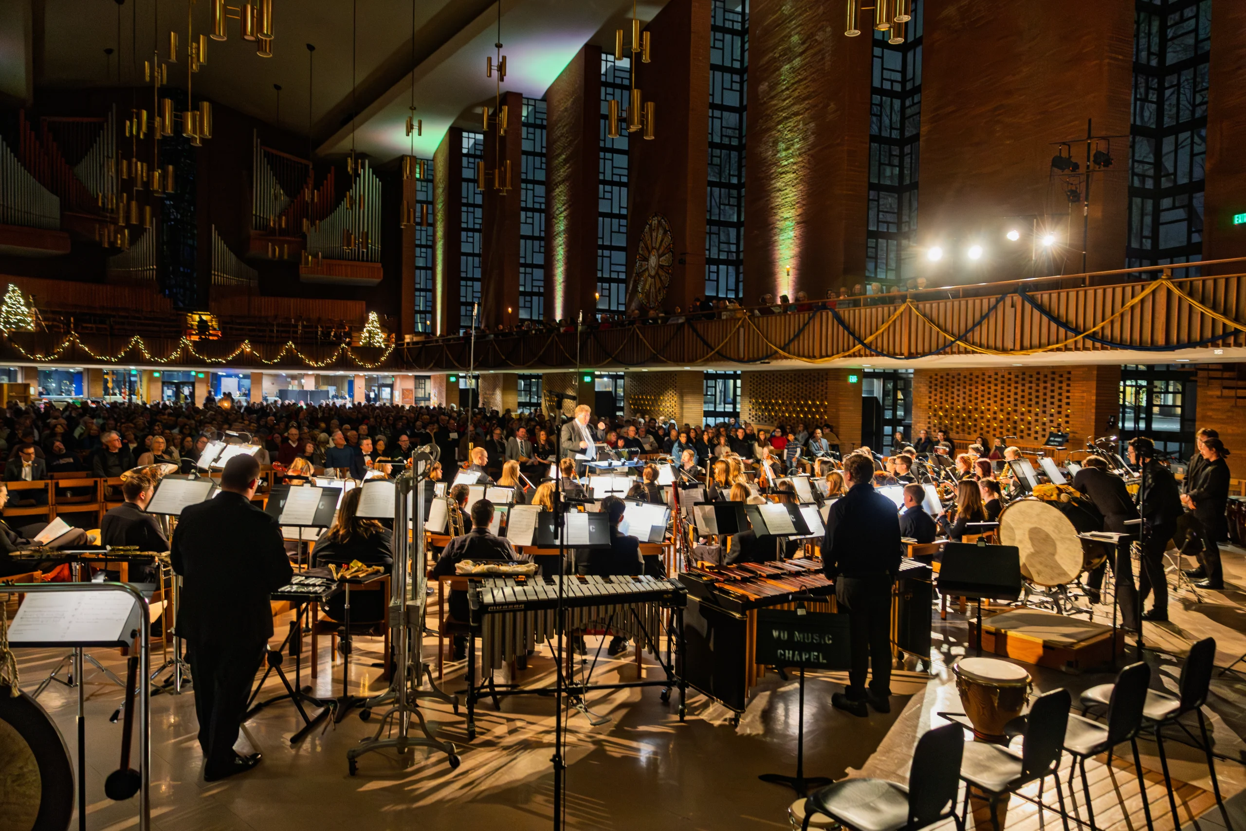 A full orchestra from the Music Department performs on stage in a large, well-lit concert hall filled with an audience.