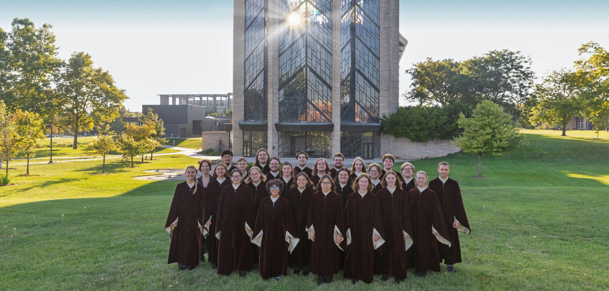 A choir in dark robes stands on grass in front of a tall, modern church with sunlight streaming through windows, evoking the spirit of Ensemble Tours.