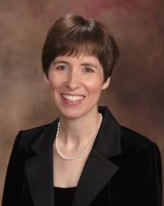 Woman with short brown hair, pearl necklace, and black blazer smiling in front of a brown studio backdrop, embodying the professional spirit of Ensemble Tours.