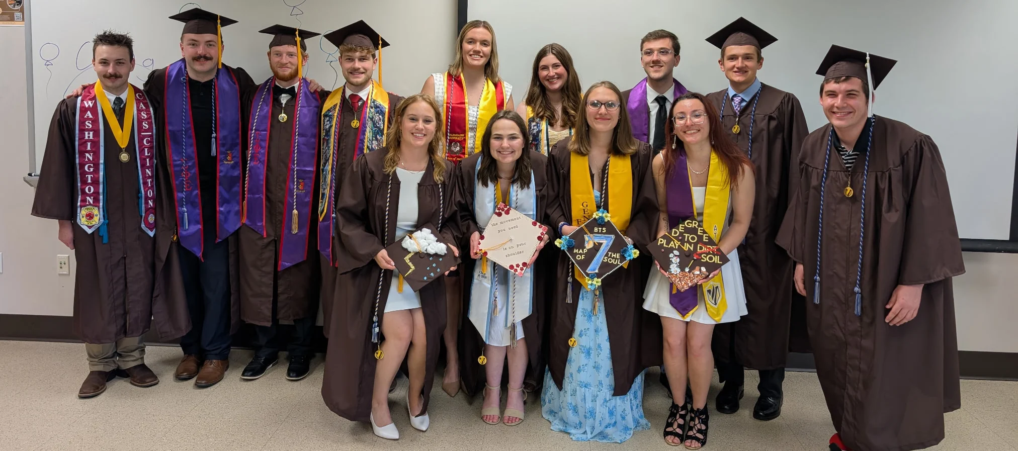 Thirteen Geography graduates in caps and gowns smile and pose indoors, some holding decorated graduation caps.