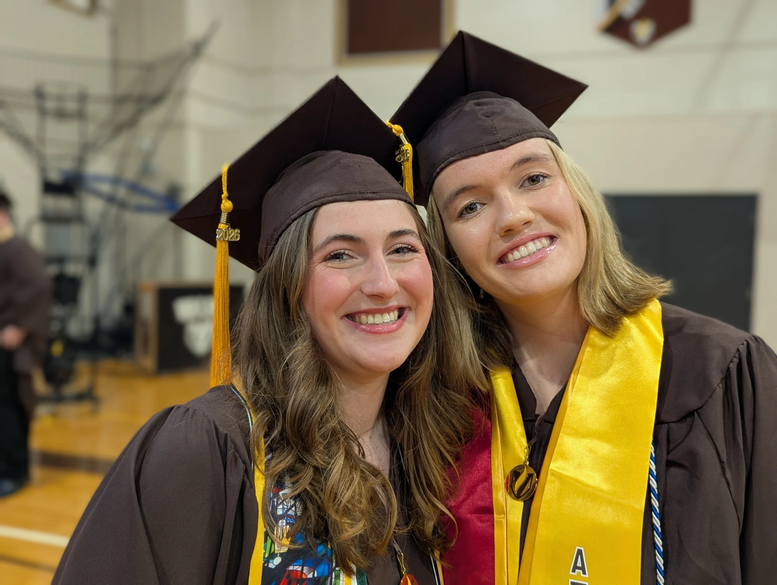 Two Geography graduates in brown caps and gowns smile together in a gymnasium, one wearing a yellow stole.