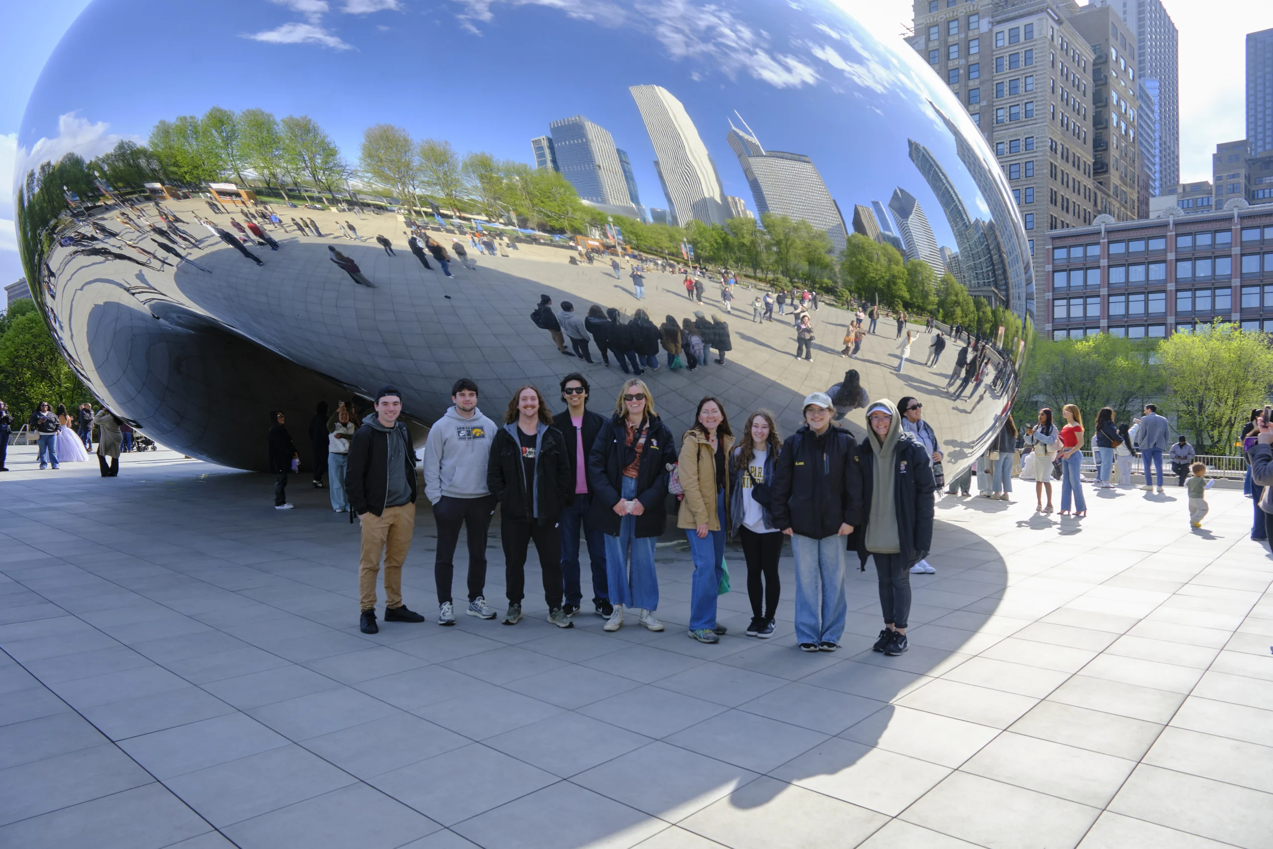 Nine people stand in front of Cloud Gate ("The Bean") in Chicago, a striking example of urban geography, with city buildings and trees reflected in its polished surface.