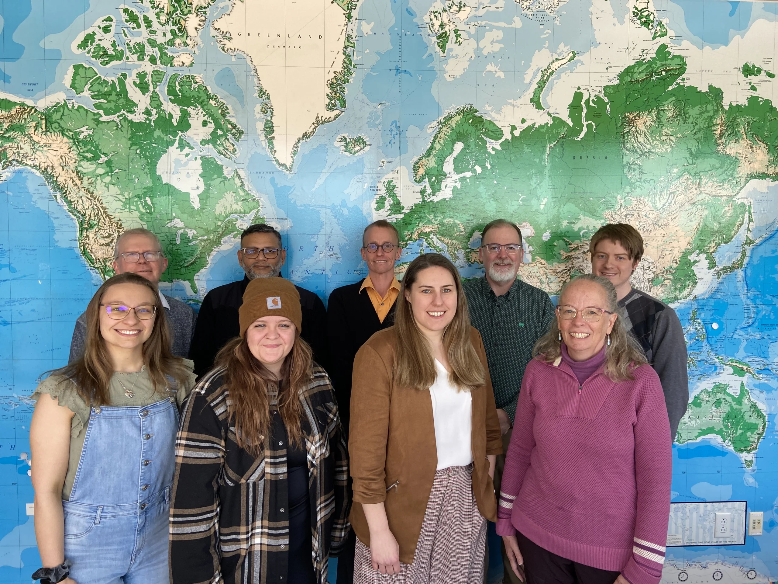 Nine people stand smiling in front of a large world map on a wall, clearly sharing their enthusiasm for geography.
