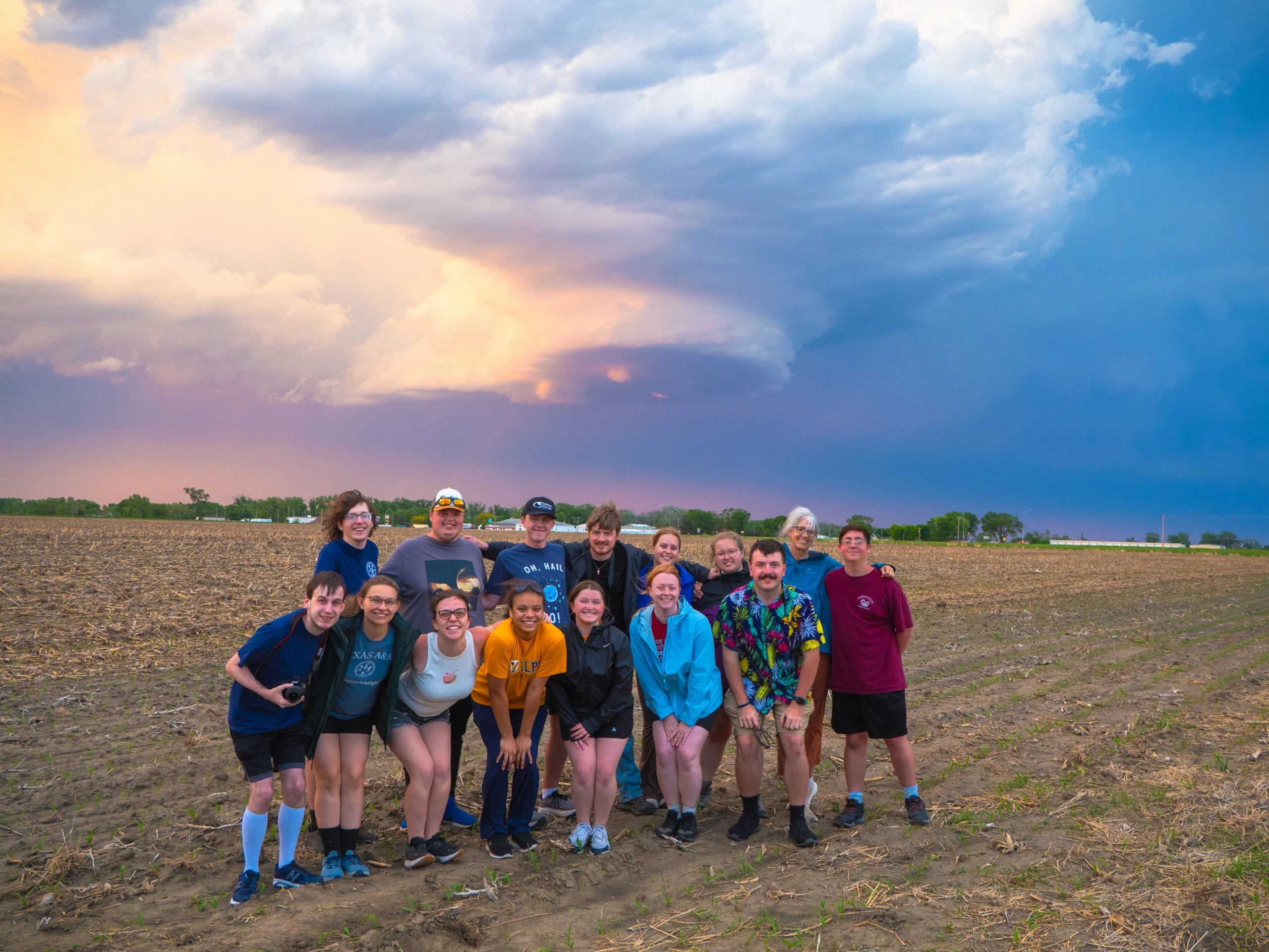 Group of people pose in a field at sunset with dramatic clouds in the sky behind them, capturing a striking moment where nature and geography beautifully come together.