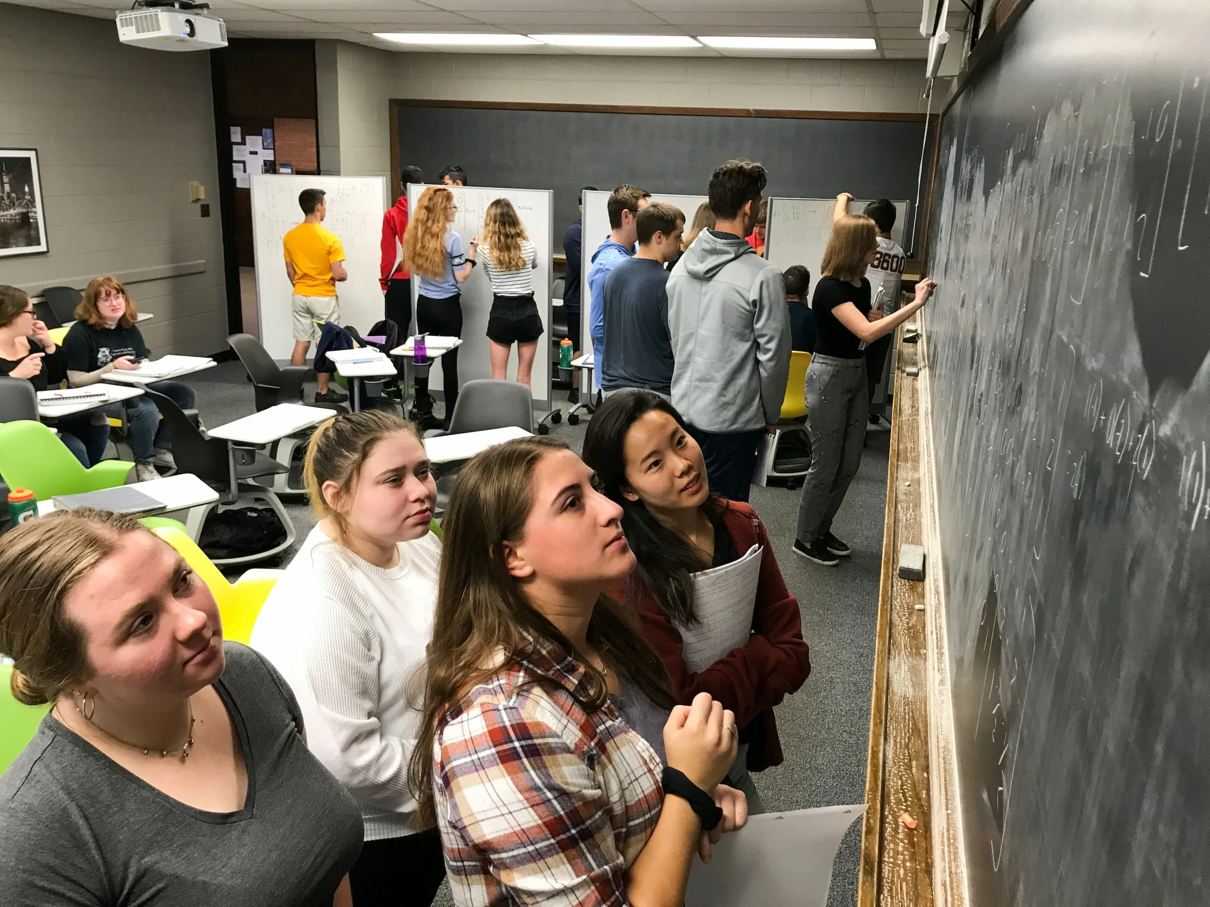 Students engage in experiential learning as they work on math problems together at a chalkboard in a classroom with others collaborating in the background.