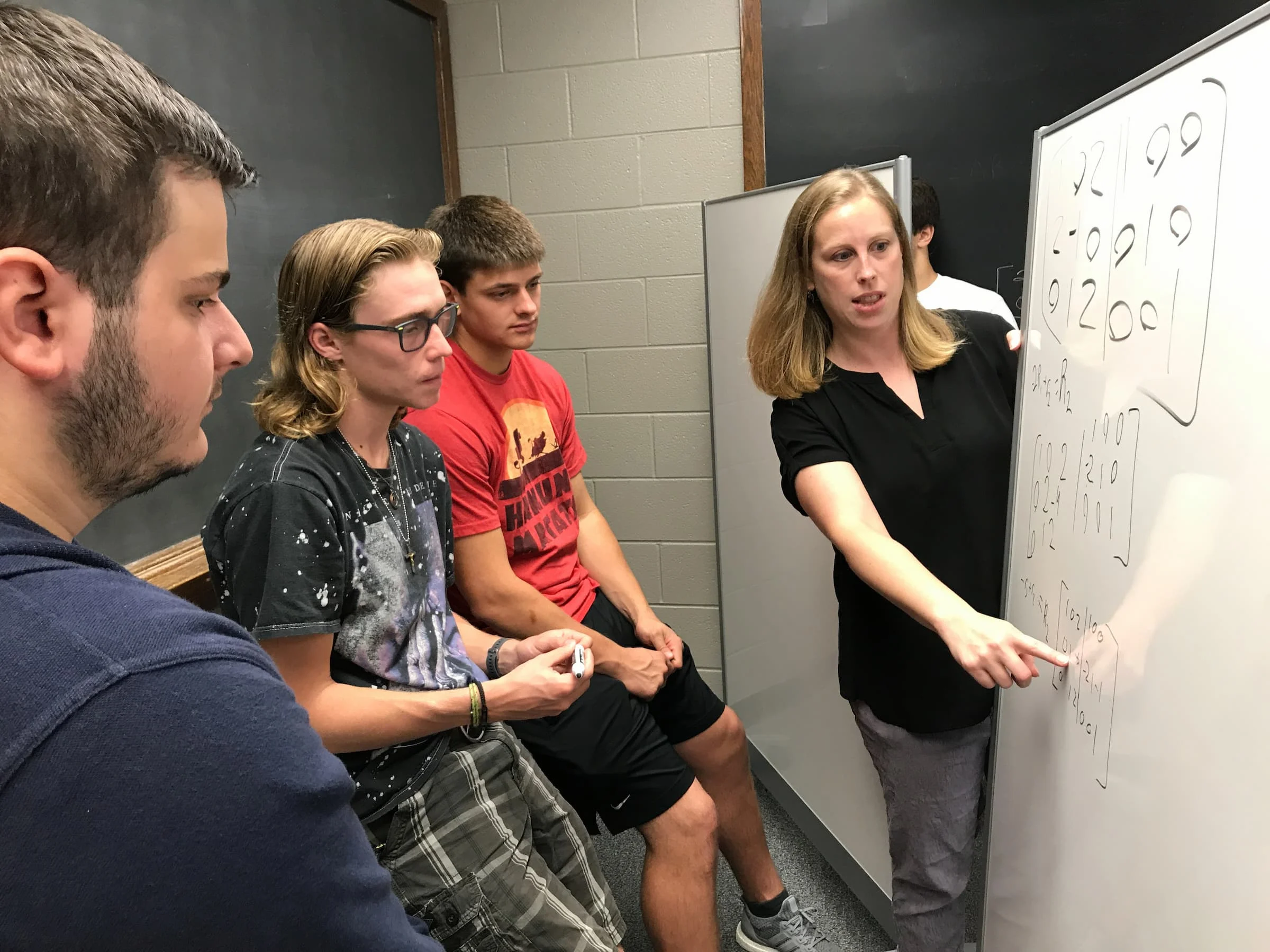A woman explains math on a whiteboard to three seated students in a classroom, integrating concepts from Information Sciences to enhance their understanding.