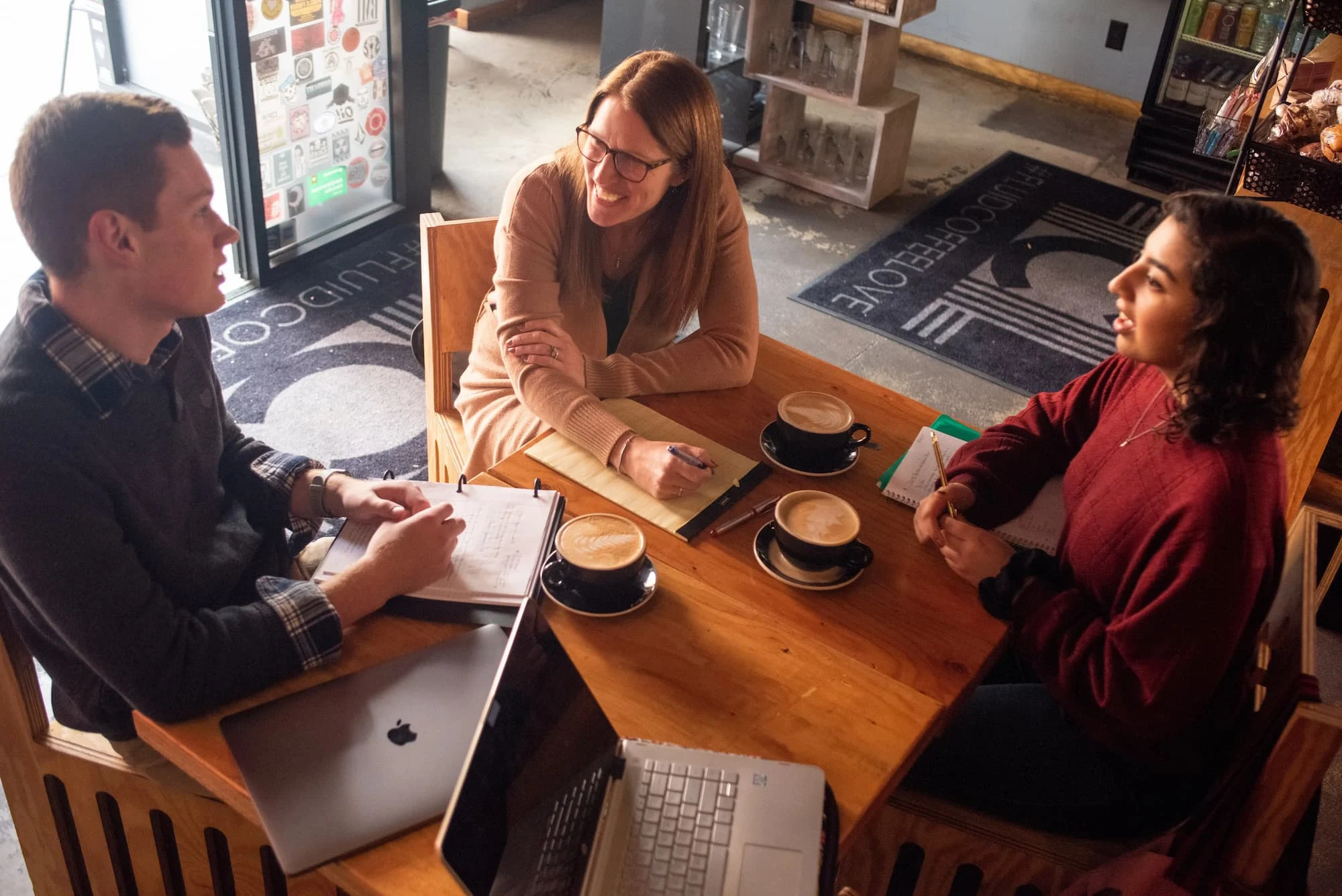 Three people sit at a café table with coffee, laptops, and notebooks, having a friendly discussion.