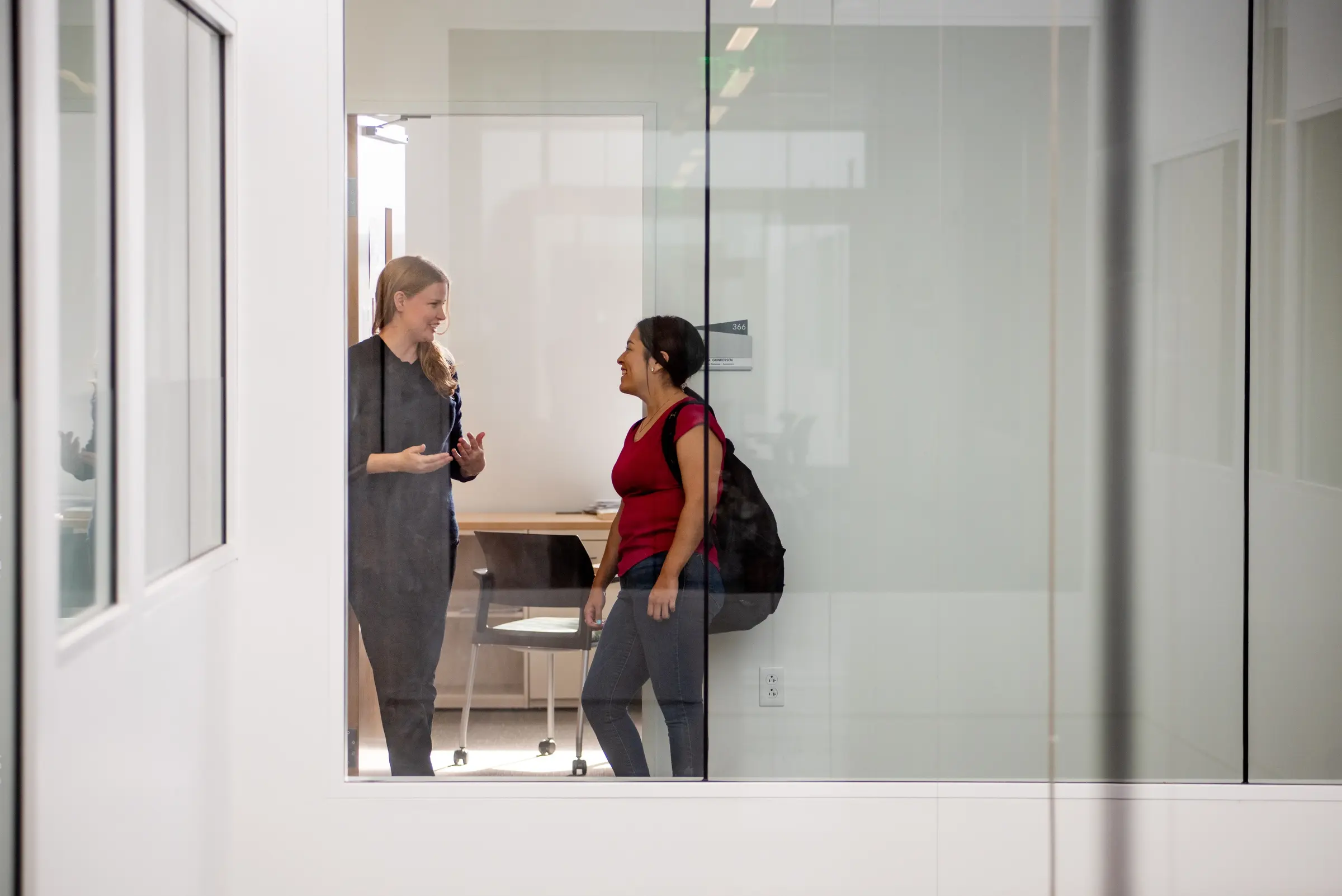 Two women stand and talk in a modern DEV office, one holding a backpack, seen through a glass wall.