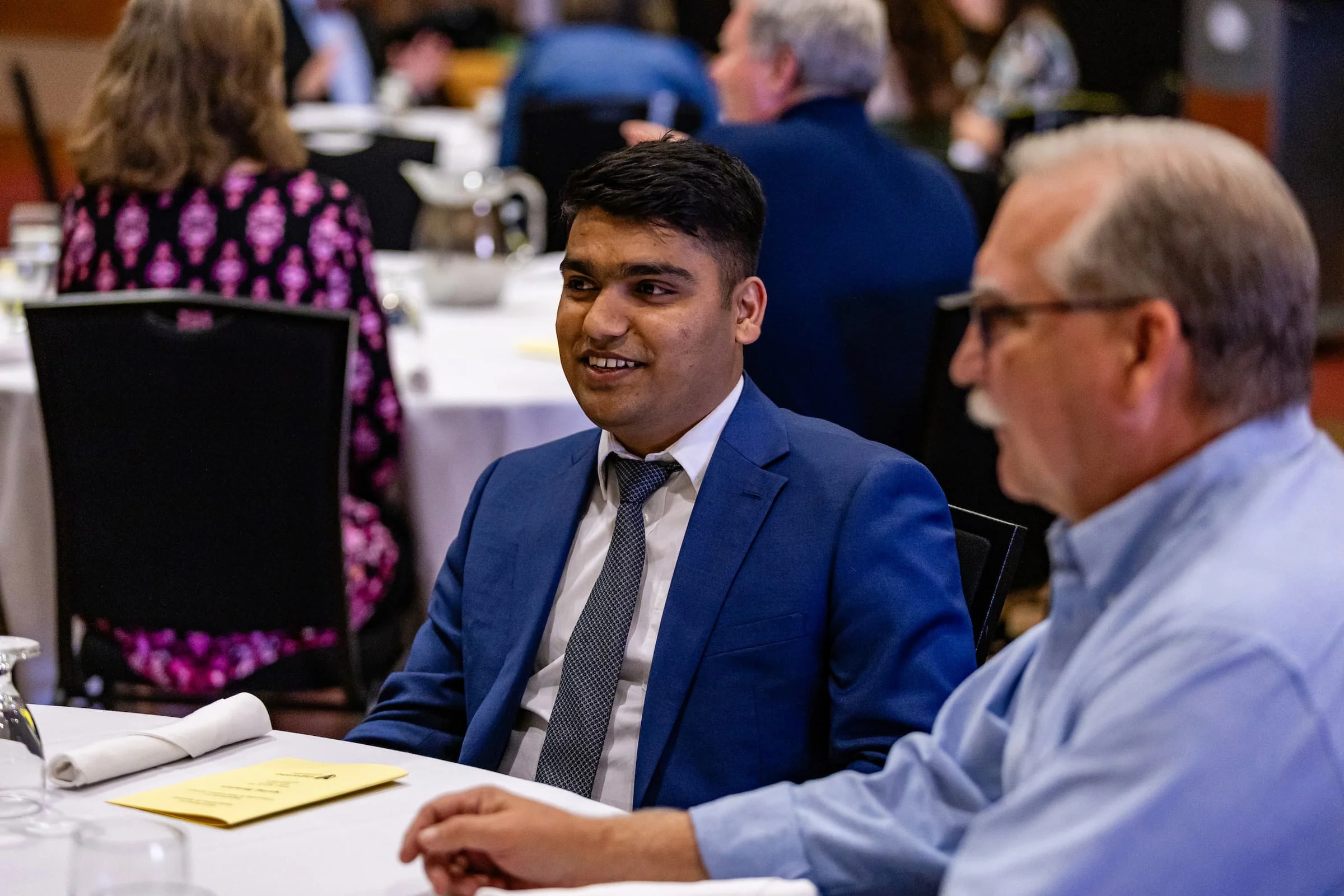 Two men in suits sit at a round table during a formal Information Sciences event, talking and smiling.