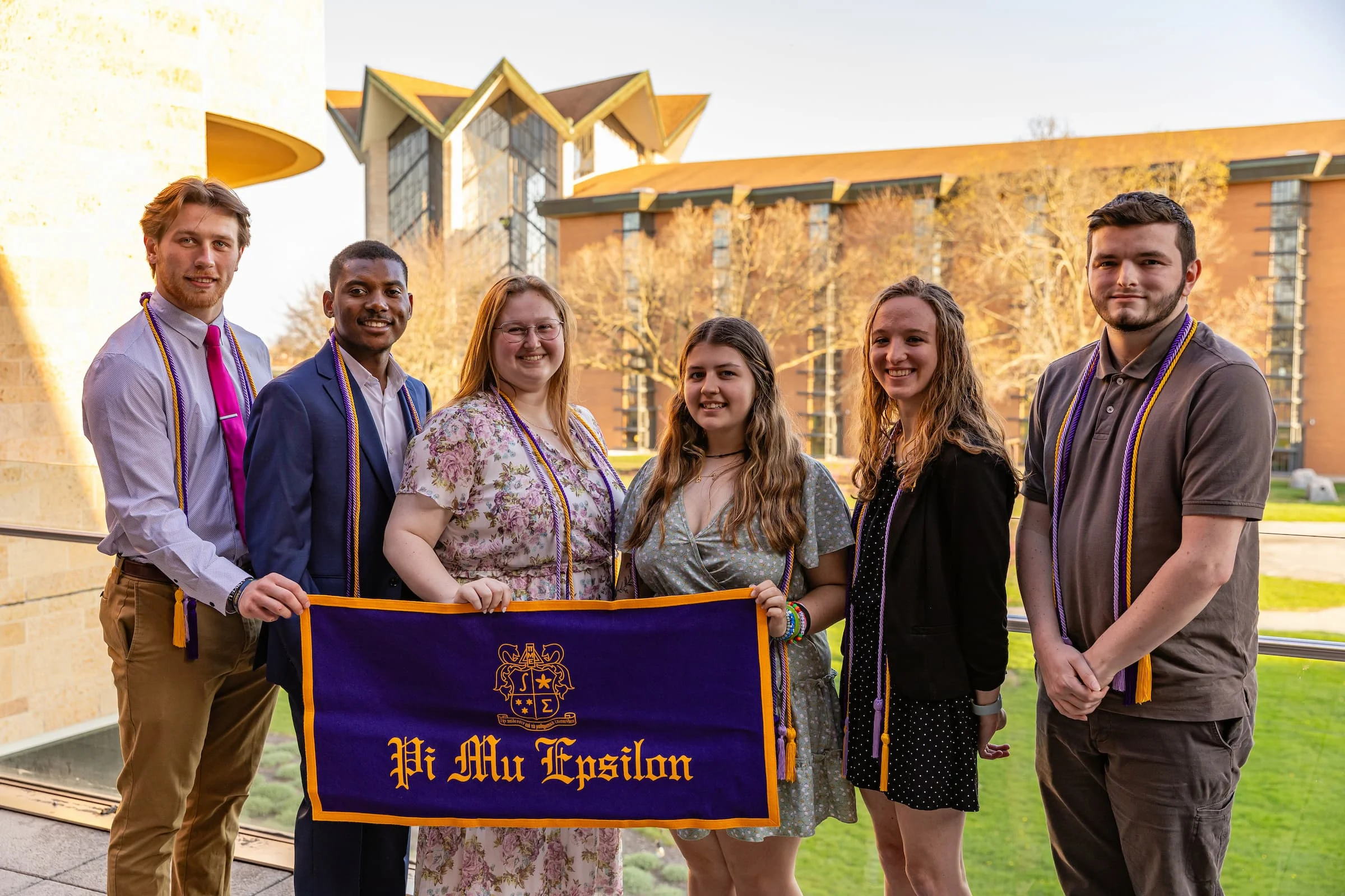Six students stand outside holding a Pi Mu Epsilon banner, proudly wearing graduation cords and smiling. Among them are Information Sciences majors, celebrating both their academic achievements and their involvement in this prestigious honor society.