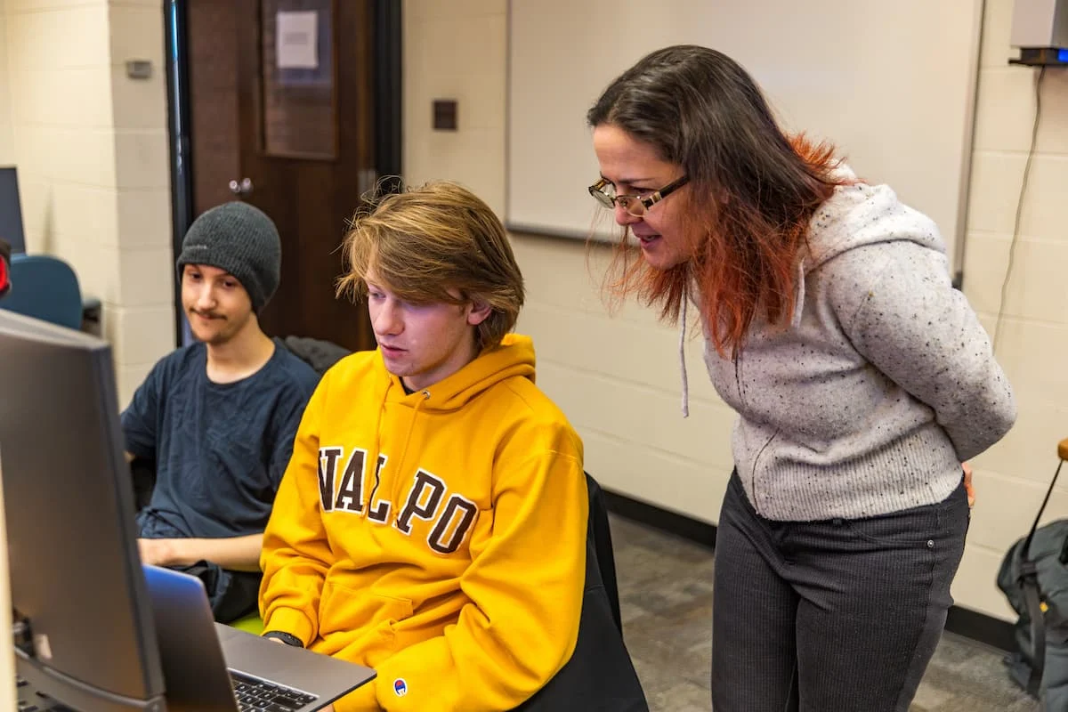 A woman stands next to two students; one in a yellow hoodie is working at a computer, engaged in an Information Sciences project.