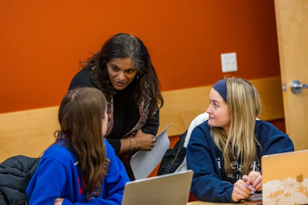 A woman stands by two students at laptops, discussing DEV tools and academic resources with them in a classroom with orange walls.