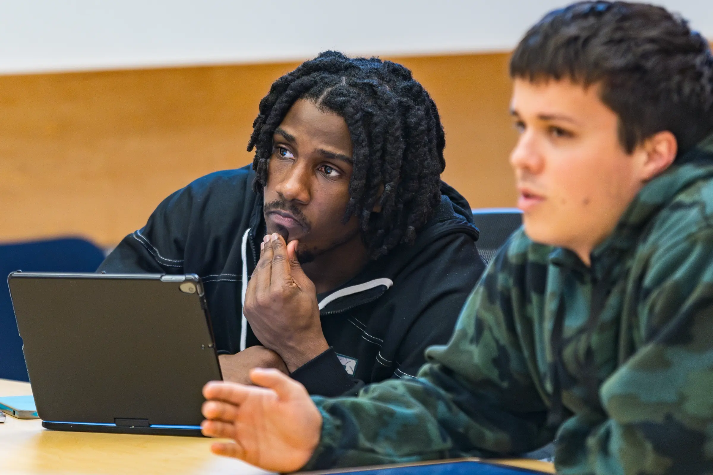 Two students in a classroom, one thinking with a tablet, the other gesturing while speaking—a dynamic scene that reflects the collaborative spirit of the School of Social Sciences.