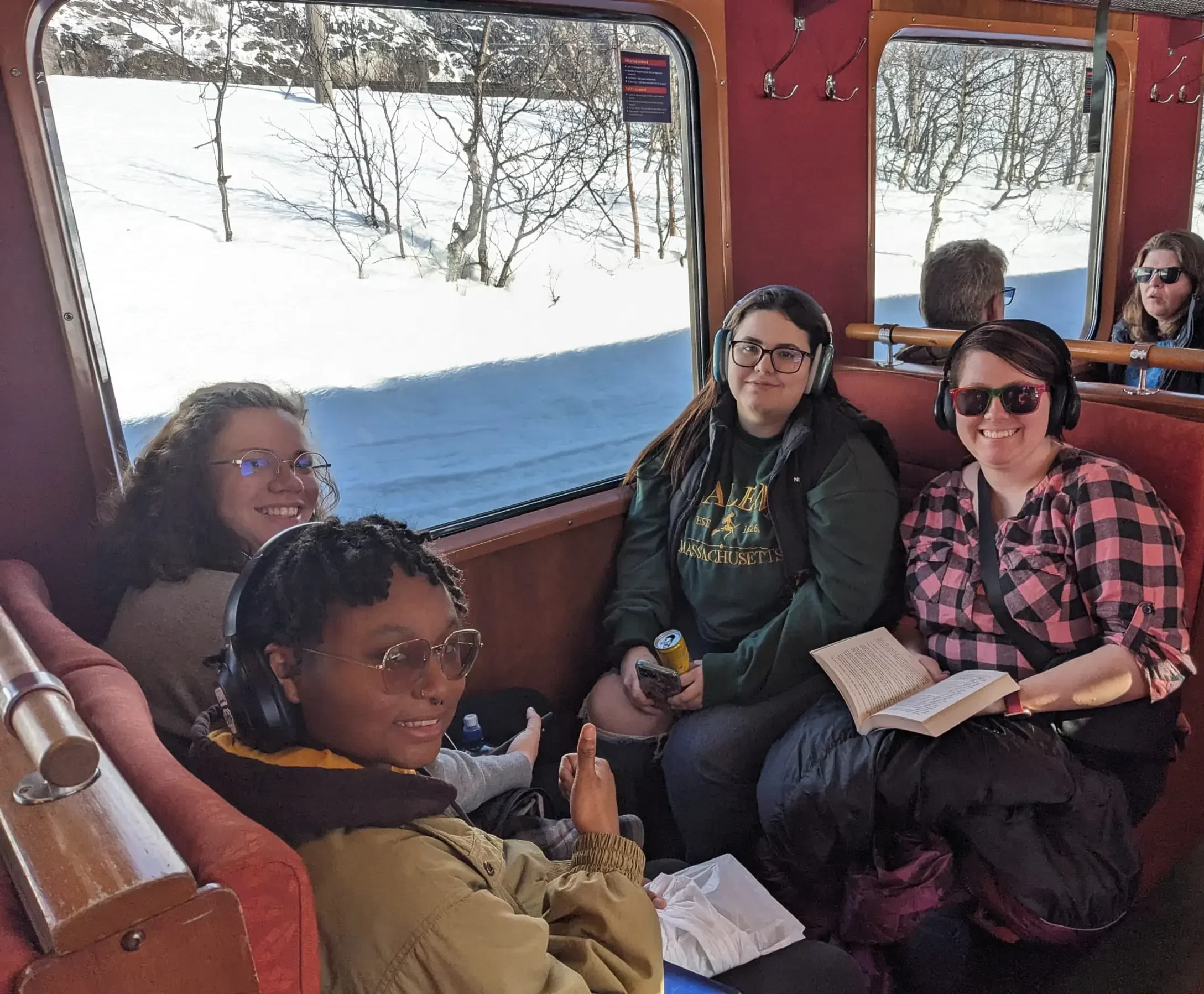 Four women sit together on a train, two with headphones, one holding a book—each absorbed in their own world of experiential learning. Outside the window, a snowy scene drifts by, turning the journey into an immersion that feels like a moving departmental overview of life and study.