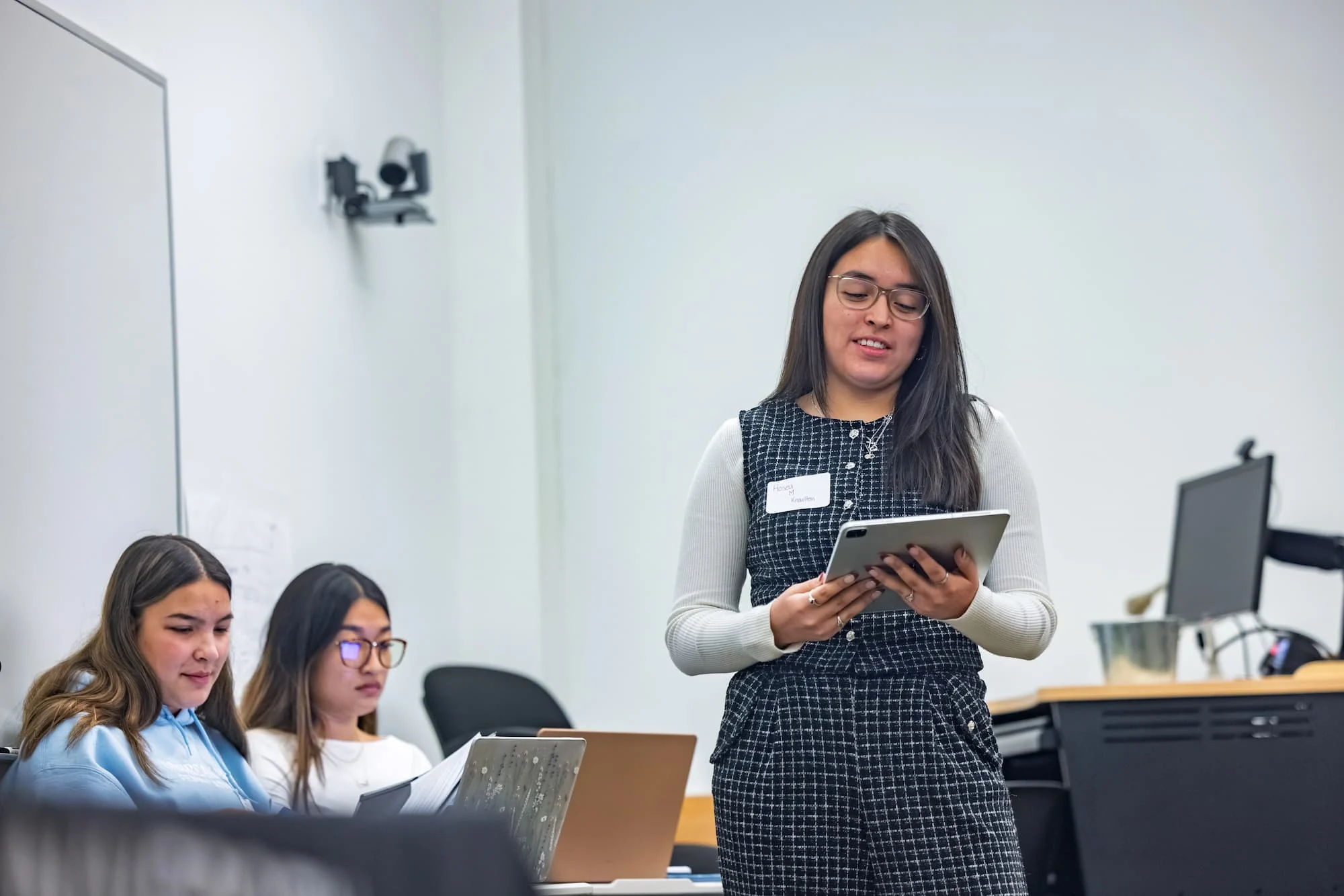 A woman stands presenting with a tablet while two others sit, working on laptops in a classroom—an engaging scene that reflects the collaborative spirit found in the School of Social Sciences.