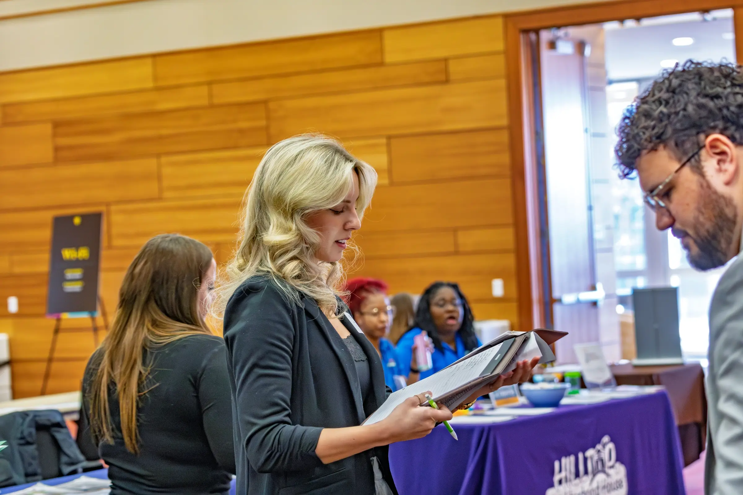 A woman holding a clipboard speaks with a man at an indoor event, where tables and people in the background contribute to an atmosphere focused on experiential learning and departmental overview.