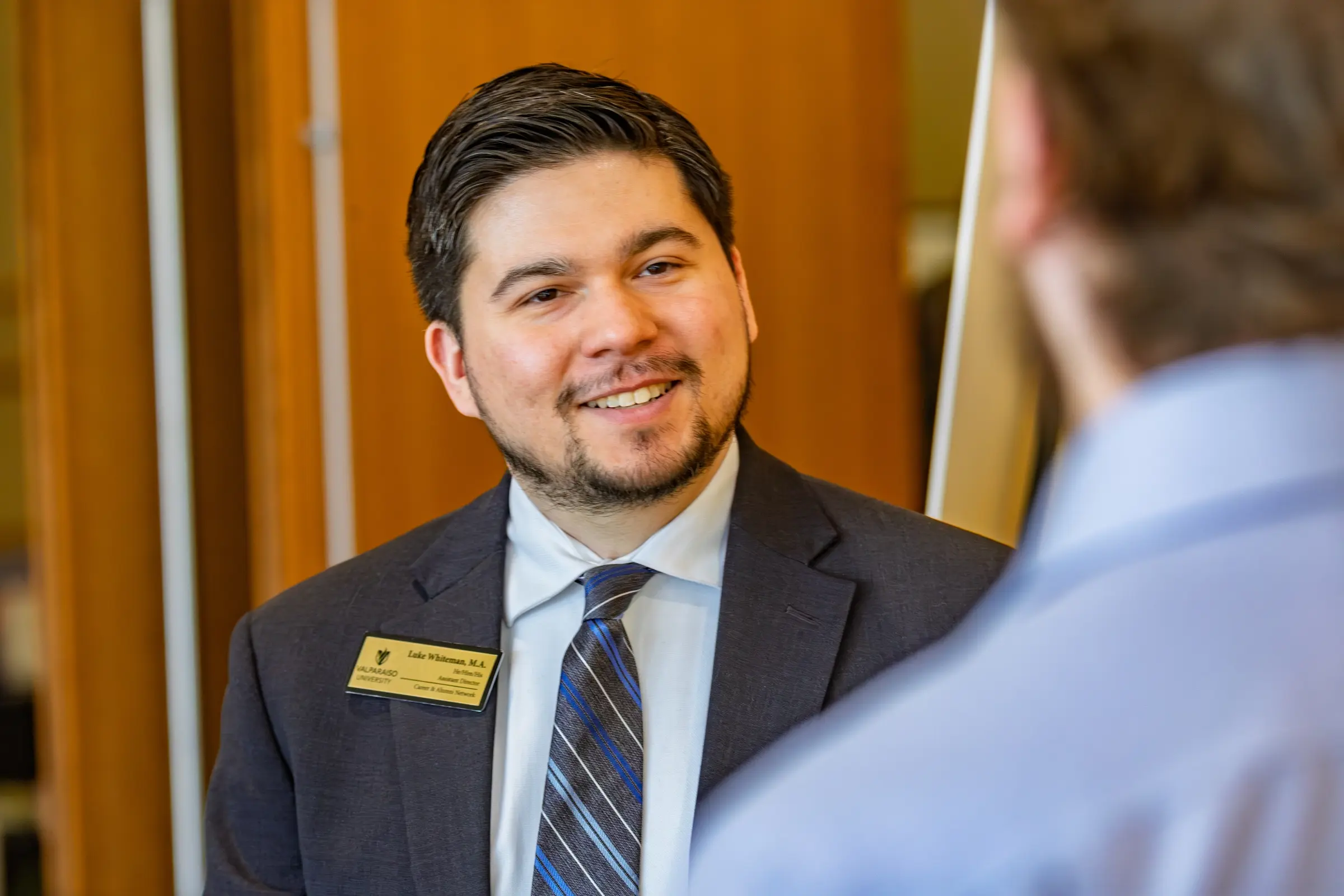 Man in a suit wearing a name badge, representing the School of Social Sciences, smiles while talking to another person indoors.