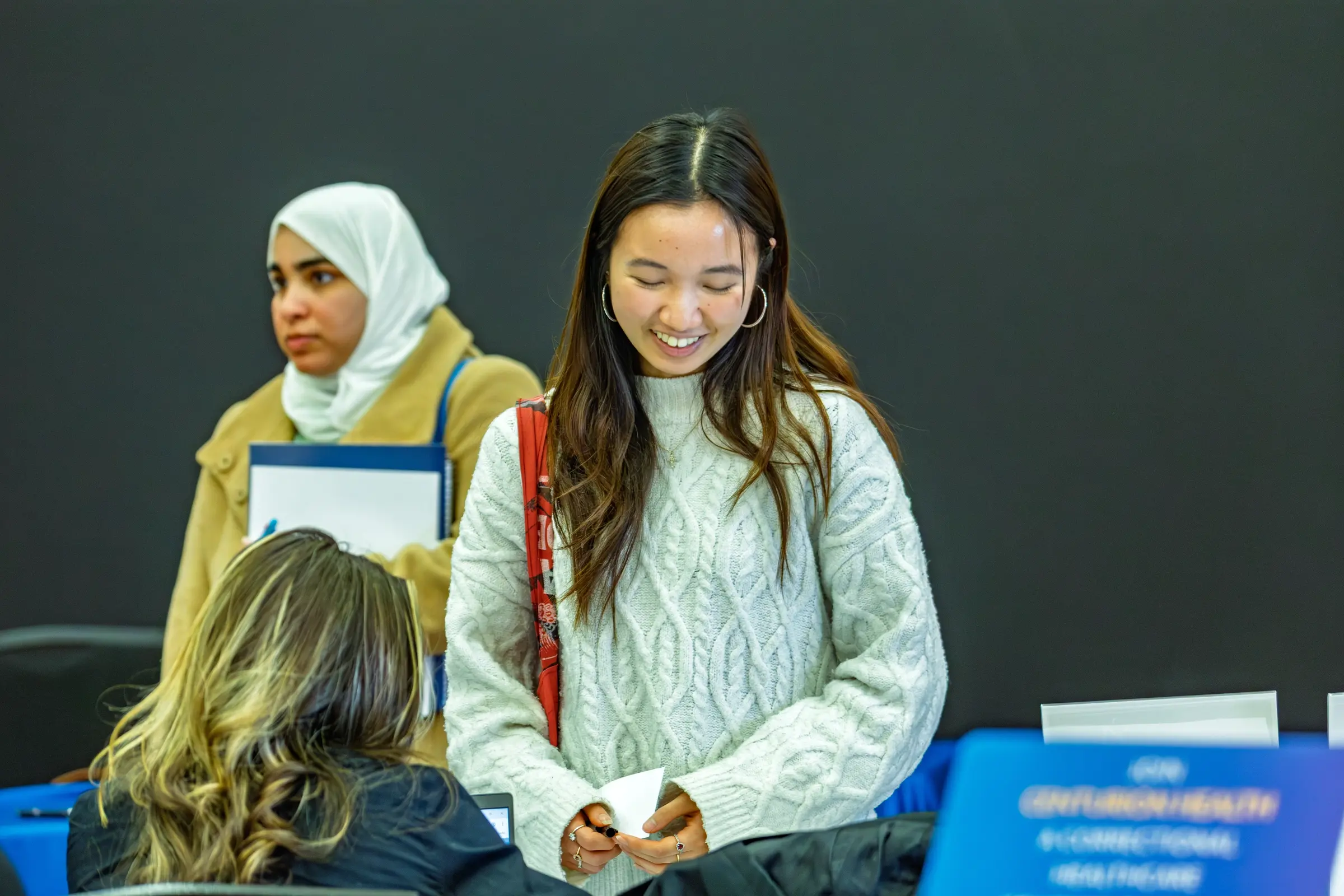 A woman in a white sweater smiles at a table, with two other women in the background at an event focused on DEV internships.
