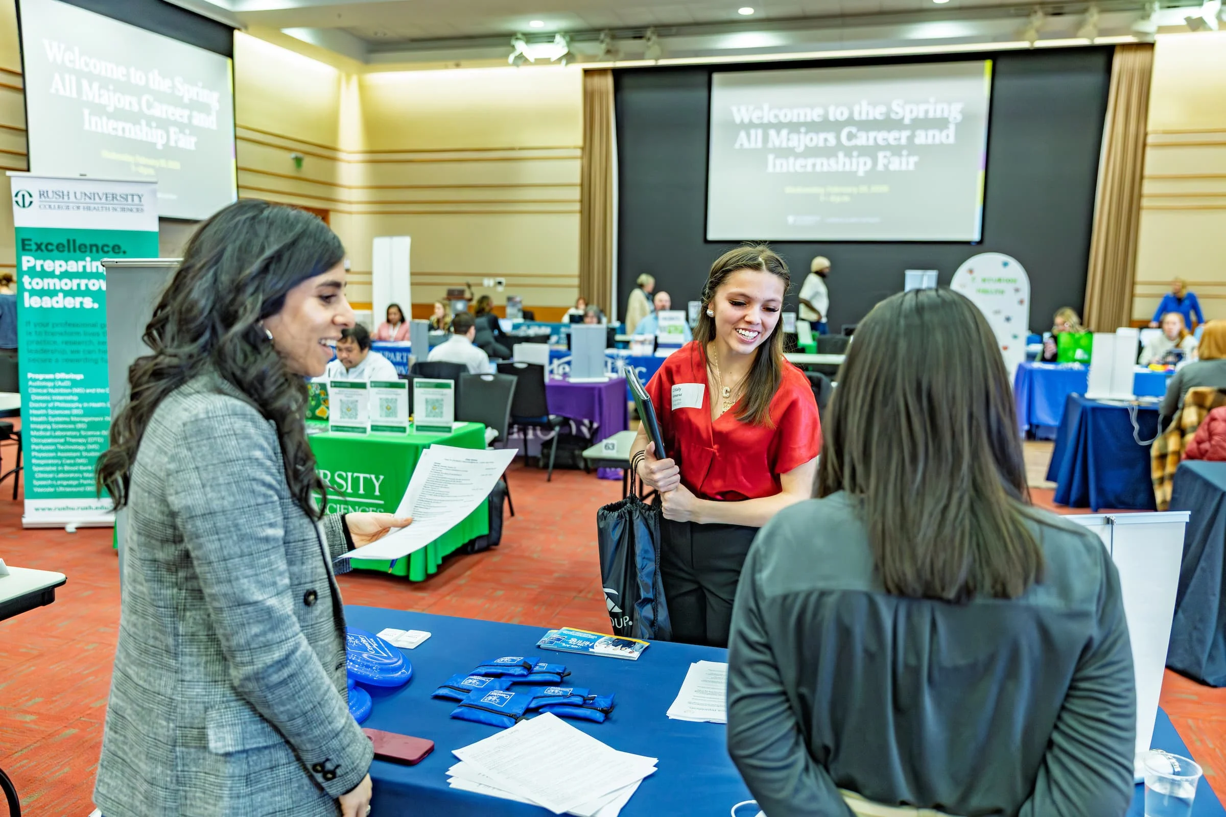 Three women talk at a career fair booth with tables, papers, and banners in a large, bright room, discussing valuable career resources and DEV opportunities.