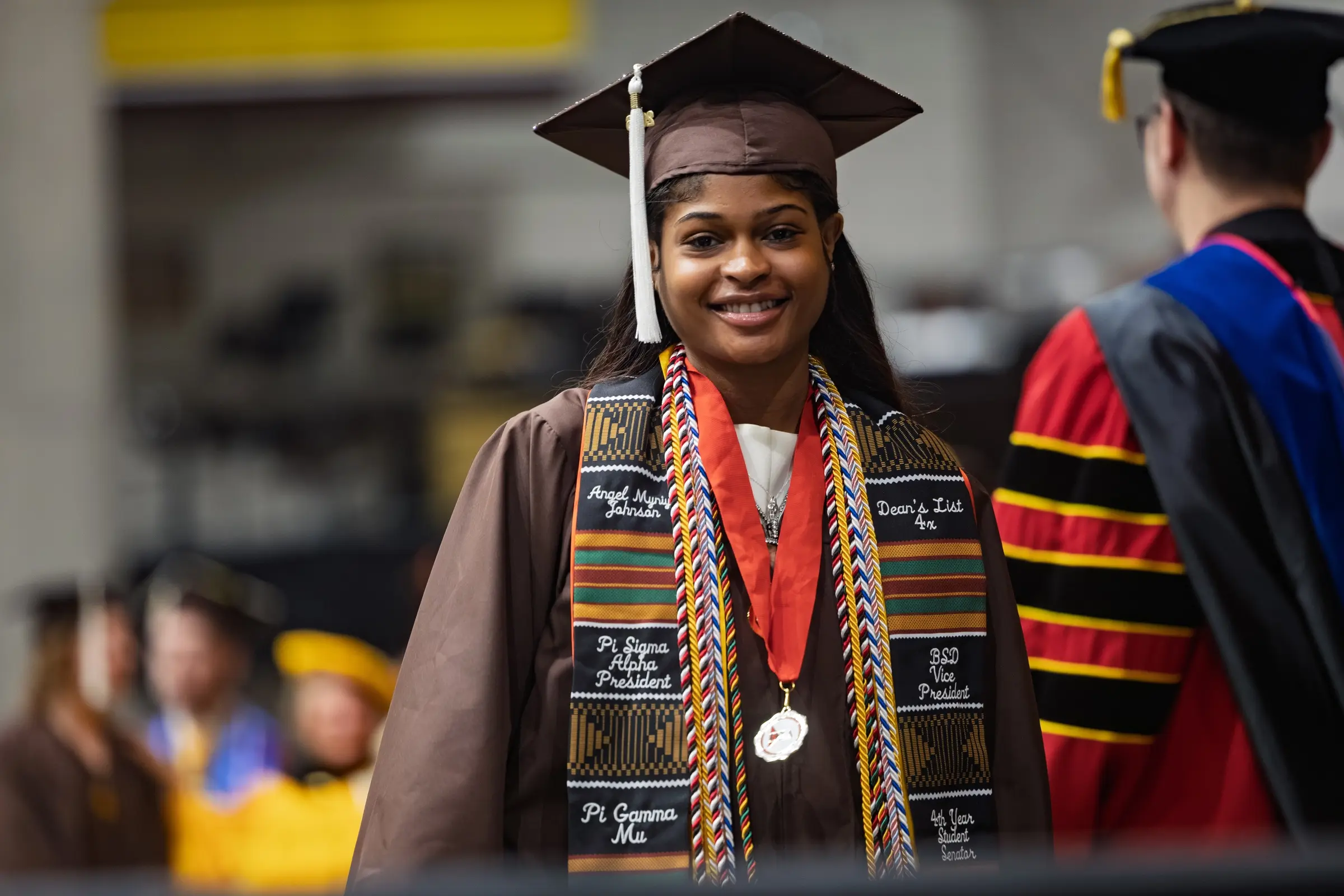 Smiling graduate in cap and gown with honor cords and stole, standing at a commencement ceremony, ready to embark on their DEV journey with the help of career resources.