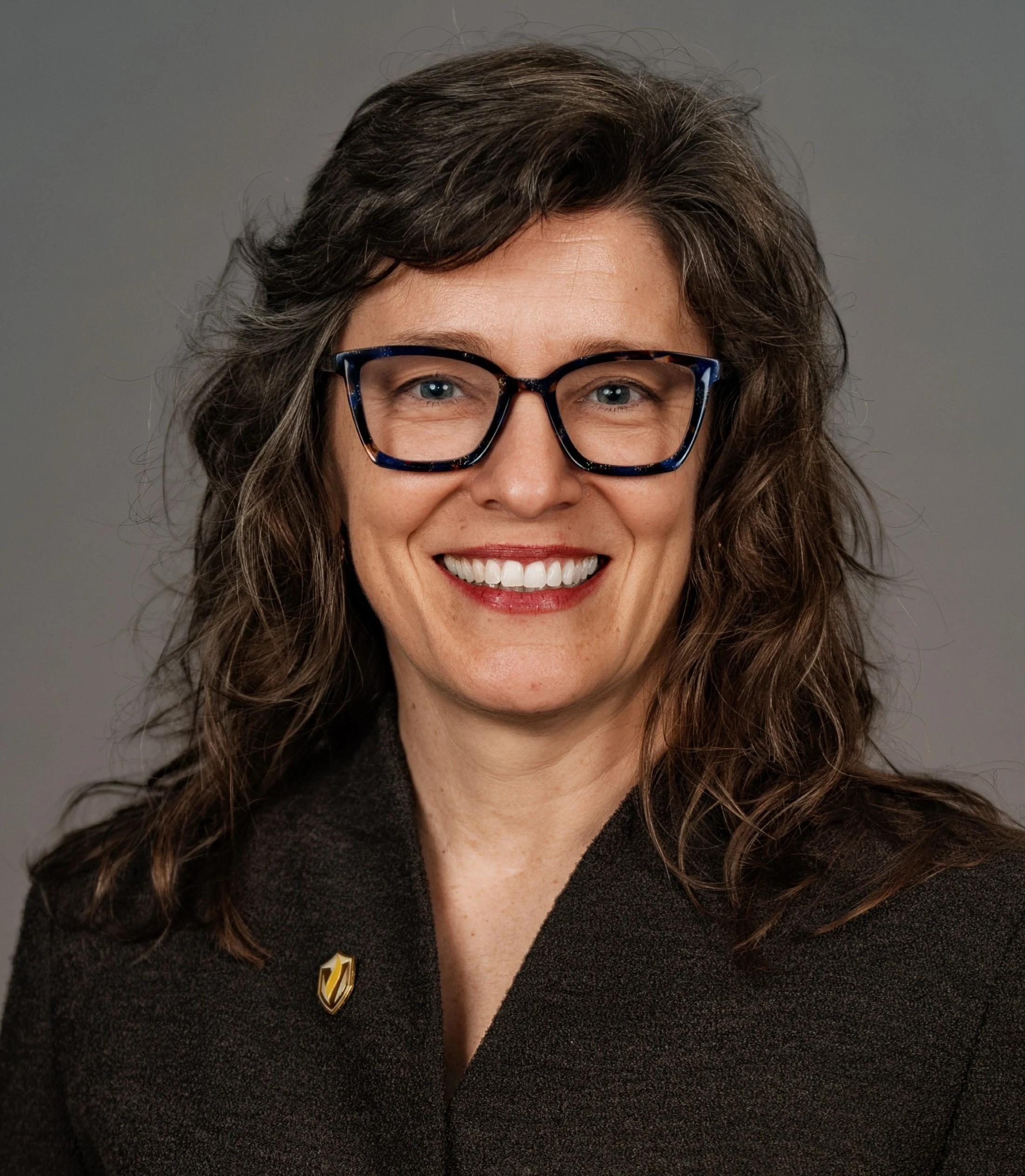 Woman with wavy brown hair, glasses, and a brown blazer smiling at the camera against a gray background, exuding confidence and leadership.
