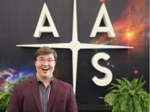 Smiling person in a maroon jacket stands in front of a large AAS sign, framed by lush plants on either side, highlighting the welcoming atmosphere for those seeking DEV and Career Resources.