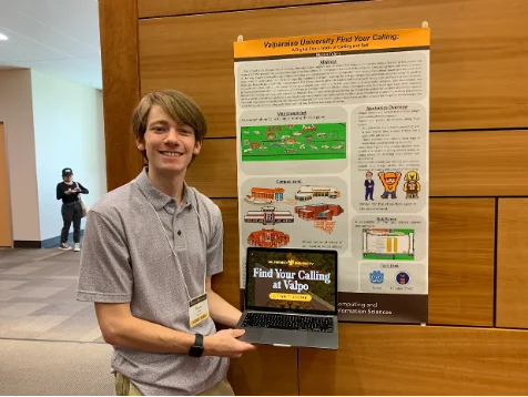 Young man smiles while holding a laptop in front of a university research poster at a conference, showcasing his DEV project and exploring new career resources.