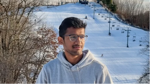 A person in glasses and a gray hoodie stands in front of a snowy ski slope with trees and ski lifts, perhaps taking a break from exploring new DEV opportunities or browsing career resources for their next adventure.
