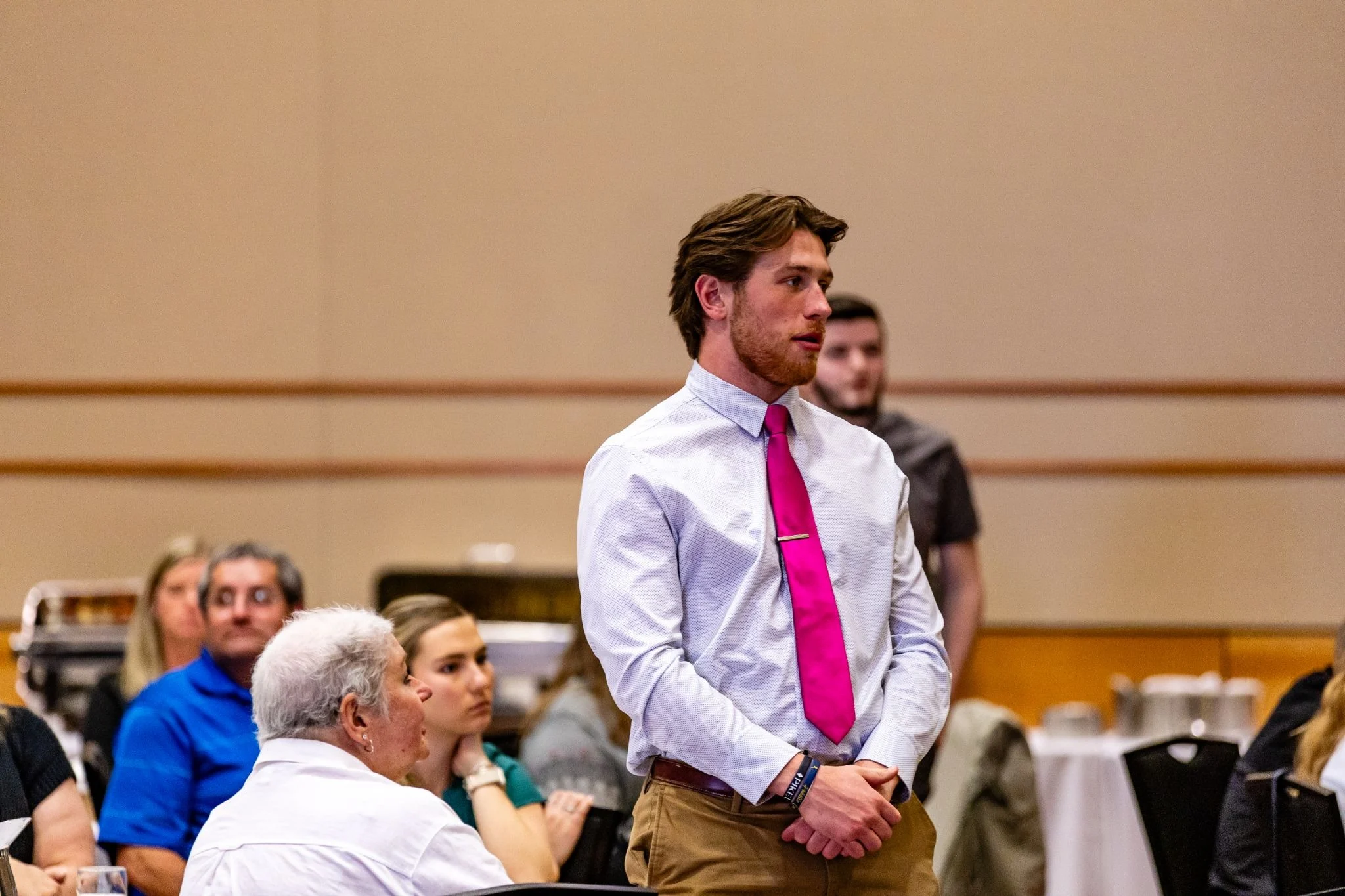 Man in white shirt and bright pink tie stands at an event focused on Experiential Learning, while others sit around tables in the background.